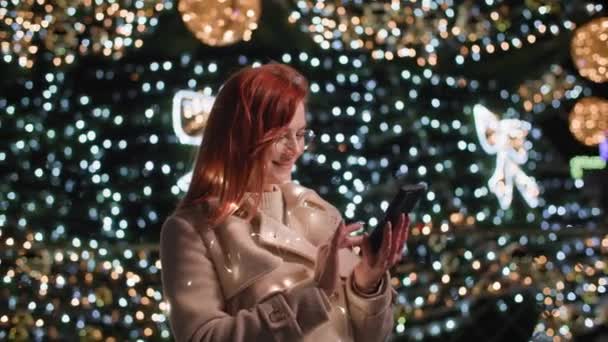 Праздничная атмосфера, молодая женщина в очках пишет поздравления, используя текстовое сообщение на фоне смартфона светящихся огней ночью в парке — стоковое видео