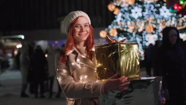 Portret van een gelukkige jonge vrouw met geschenken in haar handen wandelingen in het nachtpark achtergrond van de stad kerstboom, glimlacht en kijkt naar de camera — Stockvideo