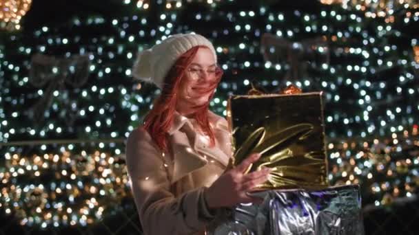 Vrolijke feestdagen, vrolijke vrouw met hoed en bril met geschenken in haar handen staat achtergrond van een feestelijke kerstboom in bloemenslingers op laat in de avond in het park, glimlacht en kijkt naar de camera — Stockvideo