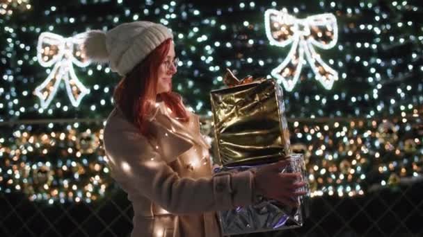 Zimowe wakacje, urocza kobieta z prezentami w rękach stoi w tle świątecznej choinki w girlandach późnym wieczorem w parku, uśmiecha się i patrzy w kamerę — Wideo stockowe