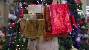 Yeni yıl satışları, elinde alışverişlerle neşeli bir kadın Noel ağacının arka planındaki perakende satış mağazasında çelenklerle eğleniyor.