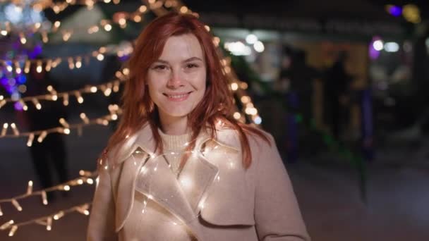Портрет красивой женщины с гирляндой на шее, улыбающейся и смотрящей в камеру на фоне огней во время отдыха в городе поздним вечером — стоковое видео