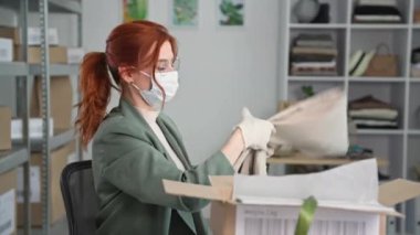 Yüzünde gözlük ve tıbbi maske olan küçük bir işletme sahibi olan genç bir kadının portresi karantinada çalışan bir online mağazada alışveriş yapanlar için kıyafet paketliyor.