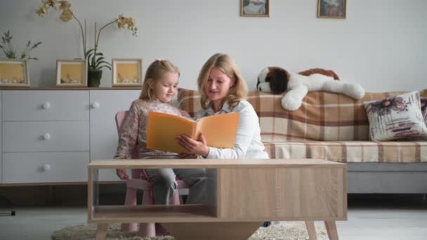 Les geven aan kinderen thuis, zorgzame vrouw met een boek in haar handen leest een boek voor aan haar dochter terwijl ze thuis zit in een gezellige kamer — Stockvideo