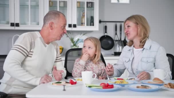 Сімейна ідилія, чарівна подружня пара зі своєю маленькою донькою весело сміються і розмовляють на кухні за столом під час обіду — стокове відео