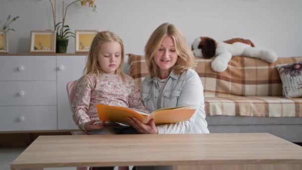 Macierzyństwo, uśmiechnięta kobieta rodzic z uroczą piękną córką czytającą ciekawą książkę siedząc przy stole w przytulnym pokoju — Wideo stockowe