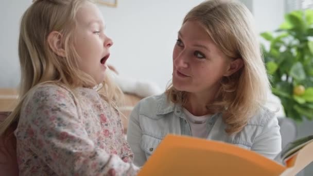 Відносини з батьками, мила дівчина читає книгу з дбайливим батьком-жінкою, чарівна дитина чхає і сміється з мамою, відпочиваючи вдома — стокове відео