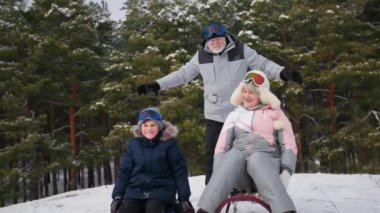 Kış tatili, torunlarıyla birlikte mutlu büyükanne ve büyükbabalar ağaçların arka planında dinlenirken karlı bir tepeden kayarak eğleniyorlar.