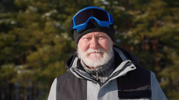 Homme séduisant âgé dans les lunettes de ski mène un mode de vie actif, se tient fond d'arbres forestiers par temps ensoleillé parmi la neige, sourit et regarde la caméra — Video