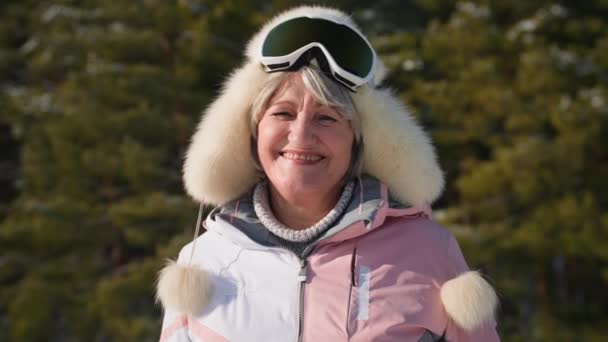 Портрет счастливой пожилой женщины в шляпе и лыжных очках ведет активный образ жизни, расслабляясь на прогулке в лесу среди снега зимой в солнечную погоду, улыбаясь и глядя в камеру — стоковое видео