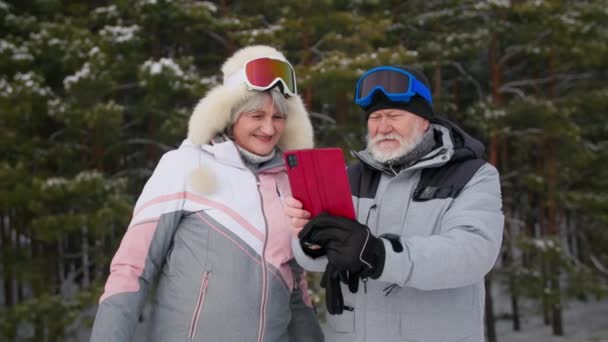 Активные пенсионеры проводят время в зимнем лесу, снимая видео на камеру смартфона на фоне деревьев — стоковое видео