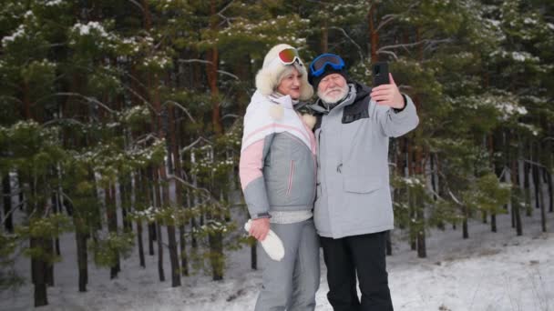 Ålderdomssemester, äldre attraktiv man och hustru kommunicera via videosamtal med hjälp av mobil kommunikation på en smartphone under en semester i en vinterskog bland snö i soligt väder — Stockvideo