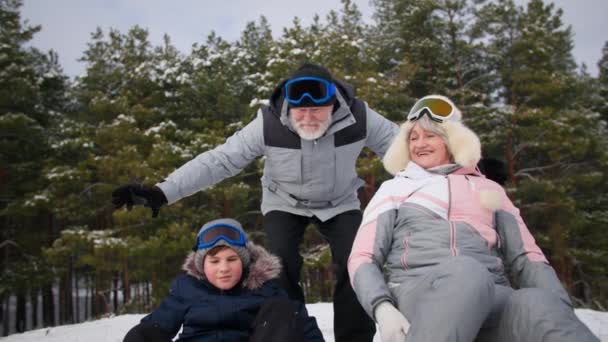 冬のアクティビティ、活発な高齢者と少年と女性は、晴れた日に森の中でリラックスしながら雪の丘から楽しいそりを持っています — ストック動画