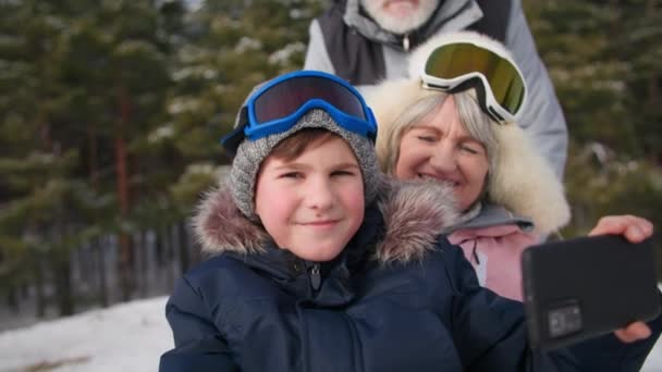 森の孫を持つ年金受給者、祖父母と一緒にスマートフォンカメラでビデオを撮影する笑顔、屋外の木々の間の雪の丘からそり — ストック動画