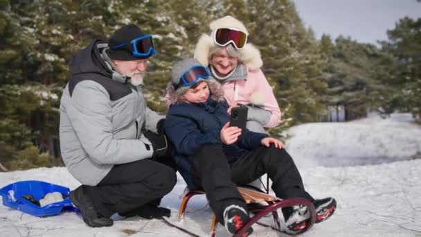 Vacances en famille, mignon enfant mâle avec femme âgée et l'homme s'amusent luge dans prairie enneigée et filmer la vidéo sur smartphone tout en se relaxant dans la forêt — Video