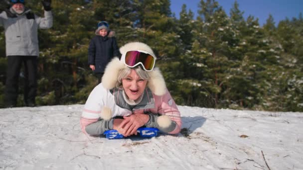 Entretenimiento para ancianos, una anciana alegre lleva un estilo de vida activo y en trineo desde una colina nevada en el fondo del bosque de árboles en tiempo soleado — Vídeo de stock