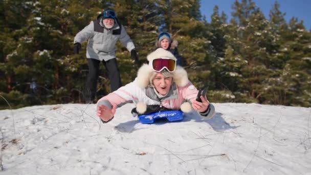 Веселые женщины на пенсии веселятся, ведя активный образ жизни катаясь на санках по снежному склону, расслабляясь с мужем и внуком в лесу в солнечный день — стоковое видео