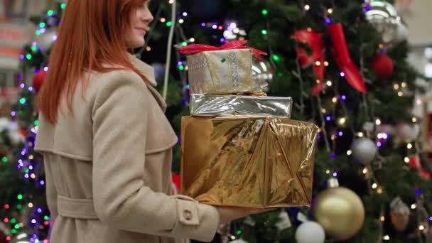 Подготовка к зимним праздникам, улыбающаяся молодая женщина с подарками в руках для семьи улыбается и смотрит в камеру на фоне елки, украшенной гирляндой в розничном магазине — стоковое видео