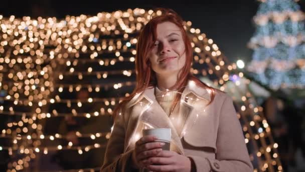 Портрет милой женщины с чашкой в руках стоит на фоне светящихся цветных гирлянд в зимний вечер в парке — стоковое видео