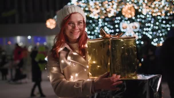 Portré boldog nő sétál ajándékokkal a kezében egy éjszakai parkban a téli szünetben, mosolyog, és belenéz a fényképezőgép hátterébe színes fények karácsonyfa — Stock videók