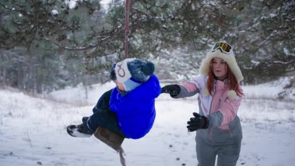 Vacaciones de invierno, feliz padre femenino rueda a su lindo hijo en un columpio cerca de un árbol en un bosque de invierno fondo de nieve — Vídeos de Stock