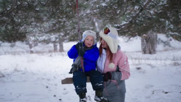 Paternidad, mujer joven cariñosa divirtiéndose con su pequeño hijo montando un columpio cerca de un árbol en un bosque de invierno en clima nevado — Vídeos de Stock