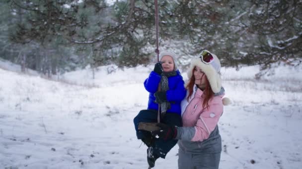 Familia diversión, adorable hijito, junto con feliz padre femenino, paseos en columpio entre los árboles telón de fondo de nieve en el bosque — Vídeos de Stock