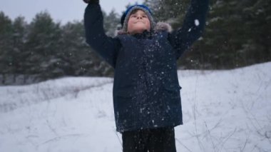 winter holidays, a happy child in hat and gloves male has fun playing snowballs in forest during snowfall background of trees
