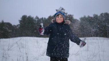 fun holidays, a charming boy throws snow and shows the class to camera while standing background of trees in winter forest