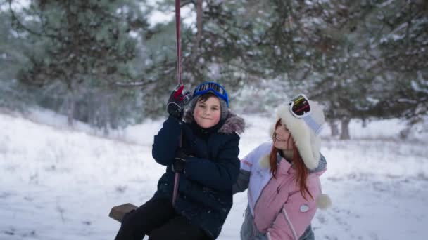 Fin de semana activo fuera de la ciudad, un hermoso niño cabalga en un columpio junto con una mujer sonriente en el bosque nevado en invierno — Vídeos de Stock