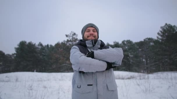 Outdoor recreation, portrait of a joyful male tourist in warm winter clothes stands background of tree in winter forest during snowfall, smiles and looks at camera — Vídeos de Stock