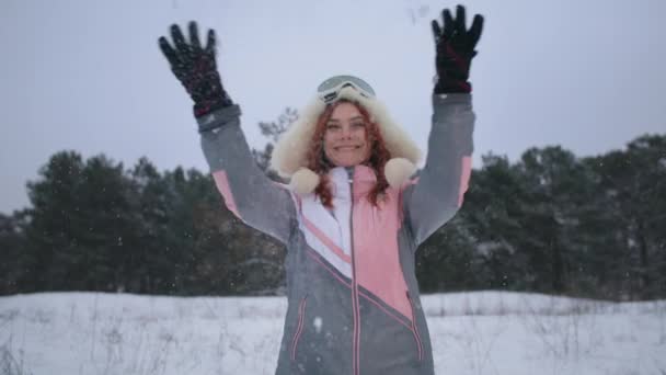Portret szczęśliwej kobiety w kapeluszu i masce narciarskiej rzuca śniegiem, uśmiecha się i patrzy w kamerę stojąc na tle zimowego lasu — Wideo stockowe