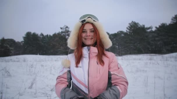 Portrait of a joyful female tourist in a hat and a ski mask throws snow, smiles and looks at camera while standing background of trees while relaxing in winter forest — Vídeo de Stock