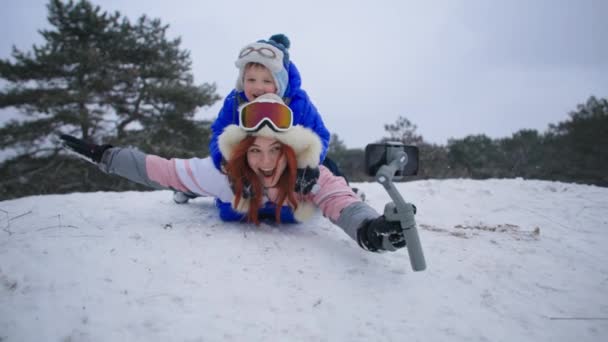 Fun winter holidays, joyful woman with her son slides down a snowy hill on a sled with a mobile phone in her hands while relaxing in forest — Stock Video