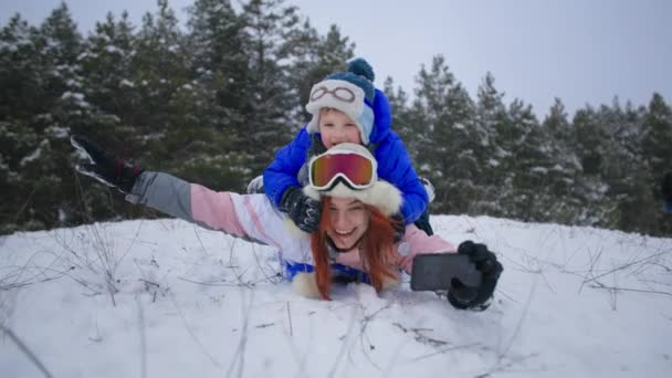 Active winter holidays, happy mother and son with a phone in their hands quickly go down snowy slope sledding in forest — Wideo stockowe