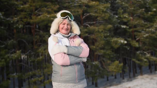 Portrét šťastné starší ženy v teplém sportovním oblečení drží ruce na hrudi při chůzi v zimním lese pozadí sněhu a stromů za slunečného počasí, usmívá se a dívá se do kamery — Stock video