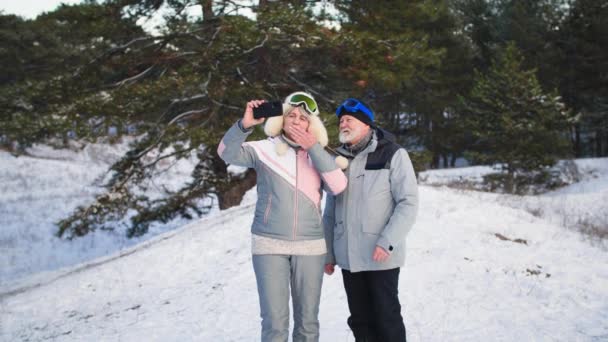 Modern glad pensionerad man och kvinna pratar video kommunikation på mobiltelefon med vänner under utomhusaktiviteter i skogen i snöigt väder på vintern — Stockvideo
