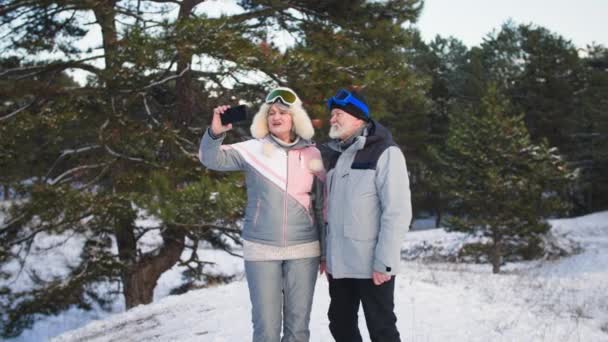 Happy pensioners lead healthy lifestyle walking in winter forest and talking with friends via video call on a smartphone background of trees among snow — Vídeo de Stock