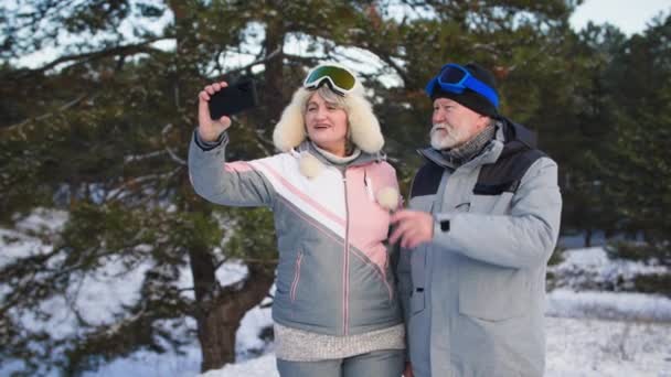 Modern gammal man med pensionerad fru pratar via videosamtal på smartphone med familjen under utomhusaktiviteter i skogen i snöigt väder på vintern — Stockvideo