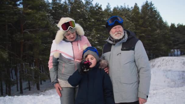 Portrait of happy elderly woman and man together with young grandson in warm clothes and ski goggles while walking in winter forest on snow background, smiling and looking at camera — Vídeo de Stock