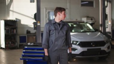 Üniformalı neşeli bir erkek çilingir portresi bir araç hizmet merkezinde bir arabanın önünde duruyor, gülümsüyor ve kameraya bakıyor.