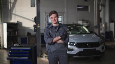 Araba servisi, elinde aletlerle üniformalı genç bir erkek tamirci arızaları teşhis etmek, gülümsemek ve kameraya bakmak için bir aracın önünde duruyor.