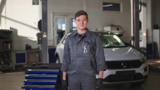 Portrait of smiling man in uniform with tools in his hands stands in front of car in vehicle service center, looking at camera — Vídeos de Stock