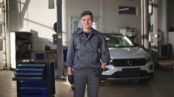 Portrait of young car mechanic in uniform with tool in his hands stands background of transport smiling and showing thumbs up, looking at camera — Stockvideo