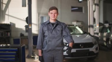 Üniformalı yakışıklı genç adam teknik istasyonda araba servisinden sonra başparmak kaldırıyor, gülümsüyor ve kameraya bakıyor.