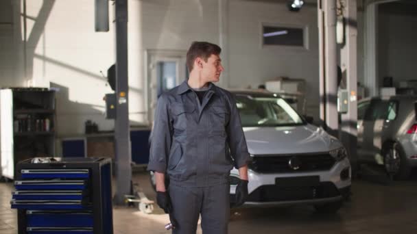 Portrait of a cute male technician in uniform with tools in his hands shows class with thumbs up standing background of car in front of vehicle diagnostics at service station — Video Stock