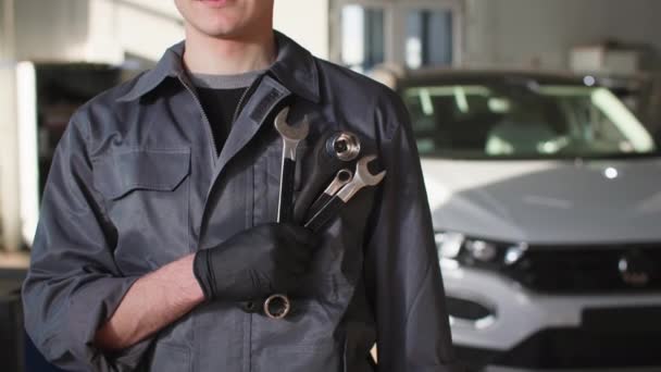 Maintenance, young professional male mechanic in uniform with adjustable wrenches stands background of car in workshop, close-up — Stock Video