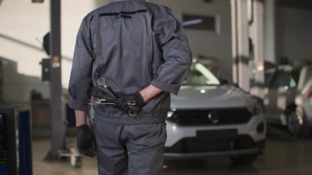 Car maintenance, a man in a uniform with tools behind his back goes to the car for repair or inspection in workshop — Wideo stockowe