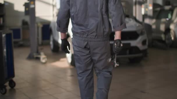 Automobile maintenance, a professional male locksmith in uniform stands with tools in his hands to inspect or repair car in service center, rear view — Stock Video