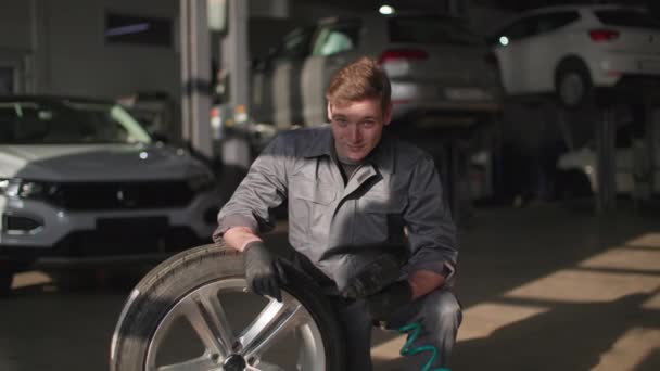 Portrait of a young male mechanic in work uniform checking a wheel in a car tire service center, smiling and looking at camera — стоковое видео